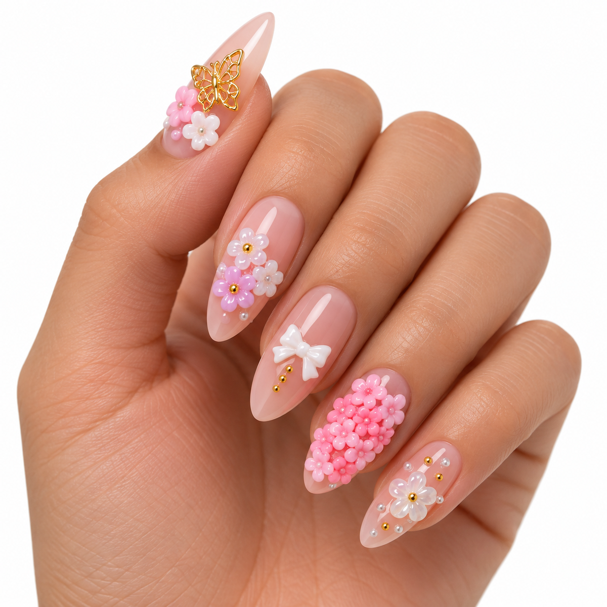 A close-up of a hand with long almond-shaped nails decorated with pink and white floral designs, 3D flower clusters, a white bow, and a gold butterfly accent on a plain white background.